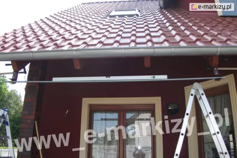 Zdjęcie dokumentuje techniczny etap montażu markizy Palladio Aluprof do krokwi dachowych budynku o ciemnoczerwonej elewacji i dachu pokrytym czerwoną dachówką. Centralnym punktem fotografii jest długa, pozioma aluminiowa rurka ułożona w gniazdach uchwytów ściennych i ułożona na niej poziomica. W ten sposób sprawdzana jest prawidłowość mocowania skrajnych uchwytów markizy. Rurka znajduje się nad dwuskrzydłowymi drzwiami tarasowymi w jasnych obramowaniach. Powyżej rurki, pod okapem dachu, widoczne są solidne, ciemne wsporniki przytwierdzone do krokwi, przygotowane do udźwignięcia ciężaru konstrukcji. Po prawej stronie kadru ustawiona jest rozstawiona, srebrna drabina aluminiowa, a po lewej stronie widoczny jest fragment drugiej drabiny, co sugeruje zaangażowanie zespołu montażowego. Budynek posiada rynny w kolorze jasnoszarym oraz okno dachowe w połaci dachu. W tle, po lewej stronie, widać fragmenty zielonych drzew i jasne niebo, co wskazuje na prowadzenie prac w pogodny dzień.