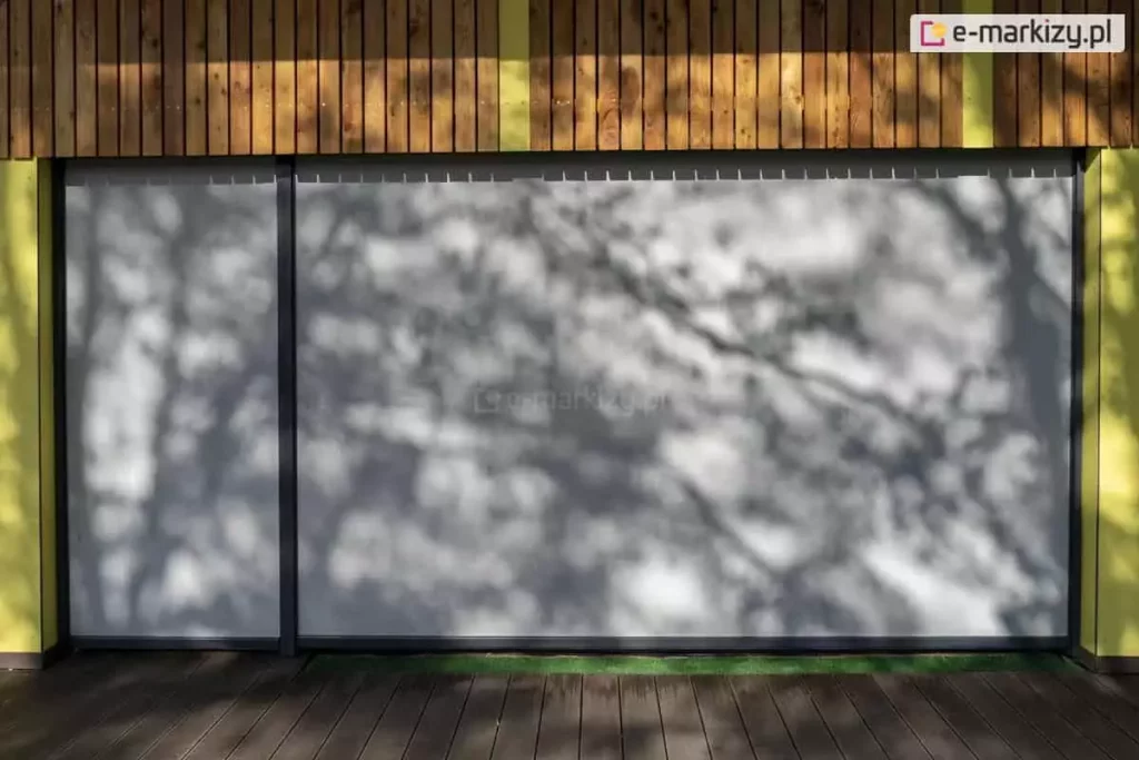 Szerokie ujęcie elewacji nowoczesnego budynku użyteczności publicznej z zainstalowanymi refleksolami Ziiip Selt. Na pierwszym planie widoczny jest drewniany taras, nad którym znajduje się wielkoformatowe przeszklenie całkowicie zasłonięte przez dwie szare rolety screen o różnej szerokości. Na napiętej tkaninie rolet malują się wyraźne, dekoracyjne cienie rzucane przez pobliskie drzewa, co podkreśla matową teksturę materiału. Górna część elewacji nad roletami wykończona jest pionowymi panelami z naturalnego drewna. Ściany boczne budynku są jaskrawo pomalowane na kolory żółty i zielony, co sugeruje, że obiekt może być placówką edukacyjną (przedszkolem lub szkołą). System osłonowy posiada dyskretne, pionowe prowadnice w kolorze antracytowym, które idealnie komponują się z nowoczesną architekturą obiektu. Zdjęcie wykonane w słoneczny dzień, prezentuje estetyczne walory rolet typu screen oraz ich zdolność do skutecznego zacieniania dużych powierzchni przeszklonych.