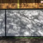 Szerokie ujęcie elewacji nowoczesnego budynku użyteczności publicznej z zainstalowanymi refleksolami Ziiip Selt. Na pierwszym planie widoczny jest drewniany taras, nad którym znajduje się wielkoformatowe przeszklenie całkowicie zasłonięte przez dwie szare rolety screen o różnej szerokości. Na napiętej tkaninie rolet malują się wyraźne, dekoracyjne cienie rzucane przez pobliskie drzewa, co podkreśla matową teksturę materiału. Górna część elewacji nad roletami wykończona jest pionowymi panelami z naturalnego drewna. Ściany boczne budynku są jaskrawo pomalowane na kolory żółty i zielony, co sugeruje, że obiekt może być placówką edukacyjną (przedszkolem lub szkołą). System osłonowy posiada dyskretne, pionowe prowadnice w kolorze antracytowym, które idealnie komponują się z nowoczesną architekturą obiektu. Zdjęcie wykonane w słoneczny dzień, prezentuje estetyczne walory rolet typu screen oraz ich zdolność do skutecznego zacieniania dużych powierzchni przeszklonych.