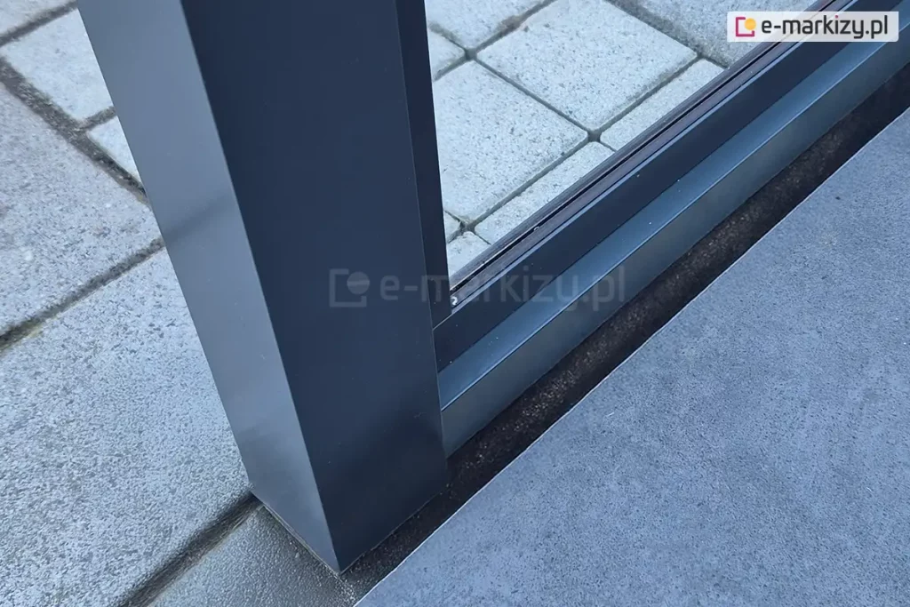Mocowanie stałej ściany szklanej Fest do zadaszenia Lunaro. Pomiędzy podporami zainstalowane zostało okno w ramie. Pod zadaszeniem widoczny taras wyłożony betonowymi płytkami i kostką.