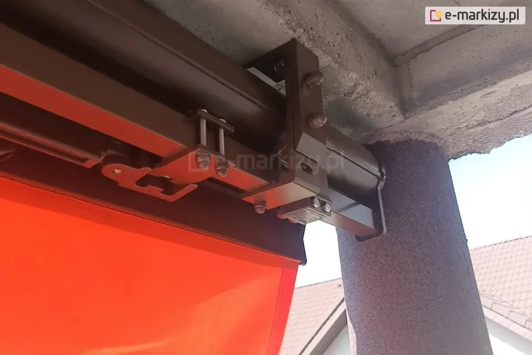 Zbliżenie na techniczne detale montażowe markizy Palladio, która została zainstalowana bezpośrednio do betonowego stropu balkonu lub tarasu. Na zdjęciu widoczna jest solidna konstrukcja wsporcza wykonana z aluminioum lakierowanego proszkowo na ciemny brąz, składająca się z masywnych uchwytów, śrub mocujących z nakrętkami oraz grubego profilu nośnego w formie rury kwadratowej. Konstrukcja jest przytwierdzona do surowej, betonowej powierzchni o porowatej strukturze, a obok widoczny jest fragment pionowego filara wykończonego tynkiem strukturalnym w odcieniu szarości. Tuz przy słupie wyraźnie widoczne jest łączenie elementu uchwytu sufitowego ze ściennym za pomocą dwóch śrub z zaślepkami. Pod kwadratową rura nośną widoczny jest fragment belki przedniej z zamocowaną w niej falbana w intensywnym pomarańczowym kolorze. W tle, pod kątem, widać fragmenty dachów sąsiednich budynków mieszkalnych pokrytych dachówką oraz jasne fragmenty elewacji pod bezchmurnym niebem.