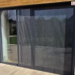 Szerokie okno tarasowe w ścianie o jasnej elewacji, nad którym znajduje się ozdobny drewniany panel, przesłonięte zostały częściowo otwartą moskitierą plisowaną Zig Zag zamocowaną do wnęki okiennej przy tarasie z dużych płyt w kolorze jasnego drewna.