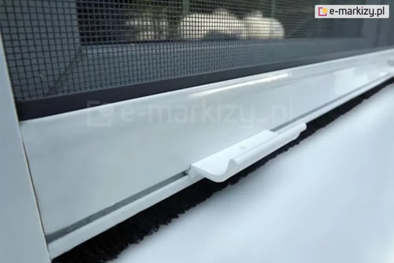 Zbliżenie białej belki dolnej moskitiery okiennej rolowanej z widocznym mocowaniem siatki, klamką ułatwiającą otwieranie i zamykanie oraz uszczelką szczotkową, zabezpieczającą przed owadami na poziomie parapetu okna.