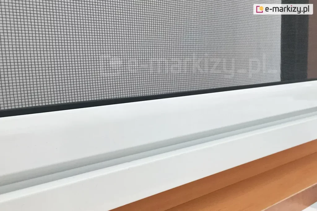 Zbliżenie belki dolnej z siatką przeciw komarom zaczepionej o prowadnicę dolną moskitiery rolowanej do okna dachowego lub tradycyjnego widoczne nad drewnianym parapetem okiennym.