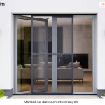 Moskitiera Slim zamontowana na dużych, przeszklonych drzwiach tarasowych, które prowadzą do nowocześnie urządzonego salonu. Zdjęcie prezentuje produkt w ujęciu od zewnątrz budynku, ukazując białą elewację z dwoma minimalistycznymi kinkietami po bokach wnęki drzwiowej oraz niską, ozdobną roślinność u dołu. Konstrukcja moskitiery Slim składa się z ciemnej, wąskiej ramy wykonanej z aluminium, która jest podzielona pionową poprzeczką na dwie równe części. Wewnątrz ramy rozpięta jest drobna siatka o wysokiej przejrzystości, która skutecznie zabezpiecza wnętrze przed owadami, nie ograniczając przy tym widoczności ani dopływu światła. Skrzydło moskitiery jest zamontowane w sposób umożliwiający jego swobodne przesuwanie.. Przez transparentną powierzchnię moskitiery Slim widać wnętrze pomieszczenia z jasną kanapą, stolikiem kawowym oraz kominkiem, w którym pali się ogień. Całość kompozycji podkreśla estetyczny i dyskretny design profilu, który idealnie komponuje się z nowoczesną stolarką otworową i minimalistyczną architekturą obiektu. W dolnej części zdjęcia na białym pasku napis "montaż na drzwiach otwieranych".