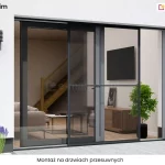 Zdjęcie prezentuje zastosowanie produktu moskitiera Slim na szerokich, dwuskrzydłowych drzwiach tarasowych o charakterze przesuwnym. Widok z zewnątrz budynku ukazuje nowoczesną bryłę o białej elewacji, na której zamontowano dwa stylowe, antracytowe kinkiety o żebrowanej konstrukcji, umieszczone symetrycznie po obu stronach wnęki okiennej. System moskitiera Slim składa się z cienkiej, niemal niewidocznej ramy w kolorze ciemnego grafitu, która przesuwa się na specjalnych szynach, chroniąc otwartą część przejścia przed insektami. Rama moskitiery posiada delikatną, poziomą poprzeczkę wzmacniającą konstrukcję, umieszczoną mniej więcej w połowie wysokości. Przez gęstą, czarną siatkę o wysokim stopniu transparentności doskonale widać luksusowe wnętrze salonu, w którym znajdują się drewniane schody z szklaną balustradą, jasna sofa, czarny telewizor na ścianie oraz niska szafka rtv z dekoracyjną rośliną doniczkową. Na tarasie, przed wejściem, po lewej stronie rosną kępy fioletowej lawendy w dekoracyjnym białym kruszywie, a po prawej widoczny jest fragment nowoczesnego, białego fotela ogrodowego o ażurowej strukturze. Całość kompozycji podkreśla techniczne atuty moskitiery Slim, takie jak minimalna grubość profili oraz możliwość integracji z dużymi przeszkleniami bez zaburzania estetyki budynku i widoku na zewnątrz. W dolnej części zdjęcia na białym pasku umieszczono napis "montaż na drzwiach przesuwnych".