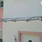 Prezentacja modułowego systemu daszków nadokiennych i naddrzwiowych Elegant zamontowanych na elewacji nowoczesnego budynku mieszkalnego. Trzy łączone moduły daszków o łukowym profilu zostały połączone liniowo, tworząc ciągłą osłonę o znacznej szerokości. Konstrukcja wsparta jest na systemowych wspornikach ze stali nierdzewnej, a wypełnienie stanowi transparentna płyta akrylowa, która zapewnia ochronę przed deszczem bez ograniczania dopływu światła do wnętrza. Daszki osłaniają białe drzwi wejściowe oraz sąsiadujące z nimi okna z szarymi roletami zewnętrznymi. Budynek posiada dwukolorową elewację: główna część wykonana jest w białym tynku, natomiast wnęki i detale architektoniczne podkreślono kolorem pastelowego różu. W tle widoczne są fragmenty sąsiedniej zabudowy z dachami pokrytymi ciemną dachówką oraz błękitne niebo, co sugeruje montaż w gęstej zabudowie podmiejskiej lub osiedlowej.
