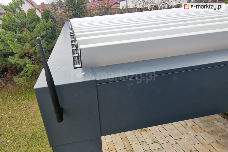 Zbliżenie na narożnik zewnętrzny pergoli tarasowej Aluprof w kolorze antracytowym, na którym zamontowana jest pionowa, czarna antena systemu sterowania radiowego. Zdjęcie prezentuje detale konstrukcyjne górnej ramy oraz ruchome lamele w kolorze białym, które są ustawione w pozycji pionowej (otwartej). Widoczny jest precyzyjny mechanizm ryglowania i łączenia poszczególnych lameli z profilem bocznym za pomocą srebrnych elementów montażowych. W tle rozpościera się widok na ogród z iglakami, trawnikiem oraz fragmentami kostki brukowej podestu. Perspektywa z góry pozwala dostrzec wykończenie górnej powierzchni profili aluminiowych oraz śruby montażowe.