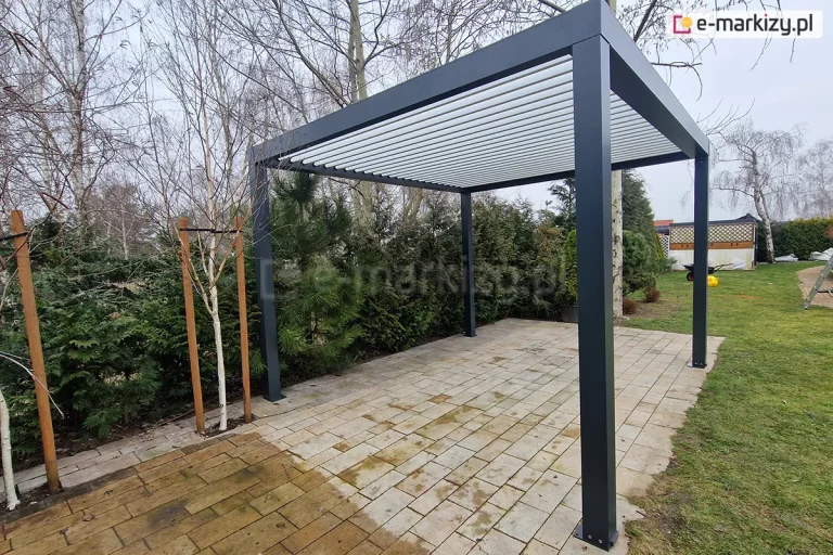 Perspektywiczny widok na wolnostojącą pergolę bioklimatyczną SB350 Selt o konstrukcji aluminiowej w kolorze antracytowym, zainstalowaną na tarasie z jasnej kostki brukowej. Dach pergoli składa się z systemu ruchomych lameli w kolorze białym, które są ustawione w pozycji uchylonej, tworząc ażurowe zadaszenie. Konstrukcja wsparta na czterech kwadratowych słupach z widocznymi stopami montażowymi. W otoczeniu pergoli znajduje się ogród z młodymi brzozami zabezpieczonymi tyczkami, gęstym żywopłotem z tui oraz fragmentem trawnika, na którym widoczna jest taczka i żółta piłka. Zdjęcie prezentuje produkt w naturalnym środowisku ogrodowym pod jasnym, zachmurzonym niebem.