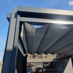 Narożnik konstrukcji pergoli wolnostojącej tako w kolorze grafitowym z rozwiniętym dachem rzymskim i widocznymi refleksolami od boku i frontu. Pod pergolą widoczny budynek i auta na parkingu. Po lewej stronie ulica z domkami i błękitne niebo.