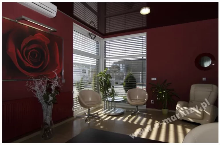 Widok na luksusowe wnętrze salonu lub poczekalni z narożnym przeszkleniem, wyposażonym w wewnętrzne żaluzje poziome fasadowe C50 Slim. Srebrne lamele są ustawione pod kątem, tworząc na jasnej podłodze charakterystyczny efekt światłocienia i zapewniając prywatność bez całkowitego odcięcia dopływu światła dziennego. Ściany pomieszczenia wykończone są w głębokim bordowym kolorze, a na jednej z nich znajduje się duża grafika przedstawiająca czerwoną różę z dedykowanym podświetleniem liniowym. Wnętrze uzupełniają dwa beżowe fotele klubowe na metalowych nogach, szklany stolik kawowy oraz nowoczesny fotel masujący. Na górze, obok klimatyzatora ściennego, widoczna jest ciemna, błyszcząca tafla napinanego sufitu, która odbija konstrukcję żaluzji i oświetlenie. Przez okna widoczny jest fragment parkingu i nowoczesnej architektury miejskiej, co podkreśla funkcję żaluzji w regulacji nasłonecznienia w mocno przeszklonych budynkach.