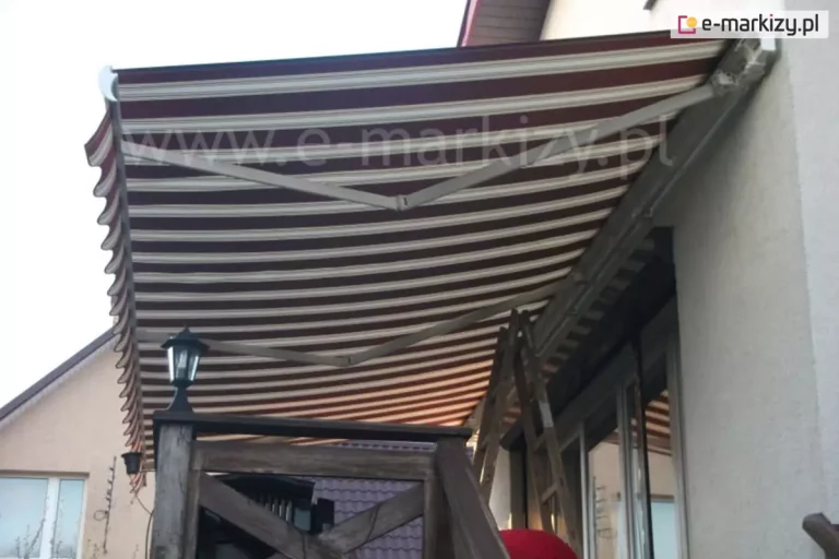 Zdjęcie prezentuje zbliżenie na konstrukcję i poszycie rozłożonej markizy tarasowej Palladio Aluprof, zamontowanej nad balkonem domu jednorodzinnego. Widok z perspektywy dolnej ukazuje dwa białe ramiona przegubowe, które podtrzymują i napinają materiał. Tkanina markizy charakteryzuje się wzorem w gęste, poziome pasy w kolorach bordowym, brązowym i beżowym. Krawędź markizy jest wykończona ozdobną, falowaną falbaną z lamówką, która zwisa swobodnie w dół. Po prawej stronie widać aluminiową kasetę przymocowaną do jasnej, tynkowanej elewacji budynku, tuż nad dużym przeszkleniem okiennym. W tle, pod markizą, stoi drewniana drabina oparta o ścianę, co sugeruje trwające prace instalacyjne lub regulacyjne. Po lewej stronie kadru widoczny jest fragment ciemnej, drewnianej balustrady balkonu z zamontowaną na niej stylową, czarną latarnią zewnętrzną ze szklanymi szybkami. W dolnej części zdjęcia widać fragment sąsiedniego budynku oraz dach pokryty ciemną dachówką. Niebo jest jasne, lekko zachmurzone, a światło wskazuje na późne popołudnie.