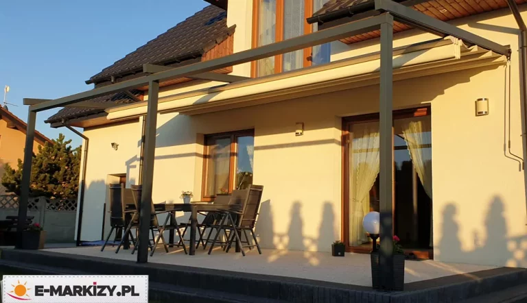 Zdjęcie prezentuje szeroką pergolę tarasową Luxo zamontowaną na ele,wacji piętrowego domu jednorodzinnego w świetle popołudniowego słońca. Konstrukcja składa się z długiej ramy aluminiowej w kolorze antracytowym, wspartej na trzech pionowych słupach o kwadratowym przekroju, które stabilizują system na podwyższonym tarasie. Zadaszenie tkaninowe w kolorze jasnokremowym jest w dużej mierze zwinięte przy ścianie budynku, co pozwala na pełną ekspozycję tarasu na słońce i uwidacznia mechanizm fałdowania materiału pod systemowym daszkiem ochronnym. Na tarasie wyłożonym jasną nawierzchnią znajduje się zestaw mebli ogrodowych z ciemnoszarymi krzesłami oraz stół, a obok słupa ustawiona jest dekoracyjna lampa ogrodowa w kształcie kuli. Cienie rzucane przez konstrukcję i barierki na elewację budynku podkreślają głębię sceny oraz stabilność całej instalacji. W tle widoczny jest fragment dachu krytego ciemną dachówką, sąsiedni budynek oraz drzewa iglaste przy ogrodzeniu.