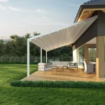 Biała pergola tkaninowa Tarasola Light w wersji przyściennej, zamontowana na jasnym drewnianym tarasie przy nowoczesnym domu o grafitowej elewacji z drewnianymi akcentami konstrukcyjnymi. Zadaszenie pergoli jest w pełni rozłożone, prezentując jasny, trójwarstwowy materiał Opatex o lekko pofalowanej strukturze, który chroni strefę wypoczynkową przed słońcem. Konstrukcja opiera się na smukłych, białych profilach aluminiowych, a pod tkaniną widoczne są poprzeczne belki zintegrowane z punktowym oświetleniem LED. Na tarasie znajduje się narożna, jasnoszara sofa z niskim stolikiem oraz osobny fotel, a w głębi, pod zadaszoną częścią budynku, widoczny jest duży stół z szarymi krzesłami ogrodowymi. Otoczenie stanowi intensywnie zielony trawnik z pasem niskich krzewów ozdobnych przy krawędzi desek oraz gęsta ściana drzew liściastych w tle, pod jasnym, popołudniowym niebem.