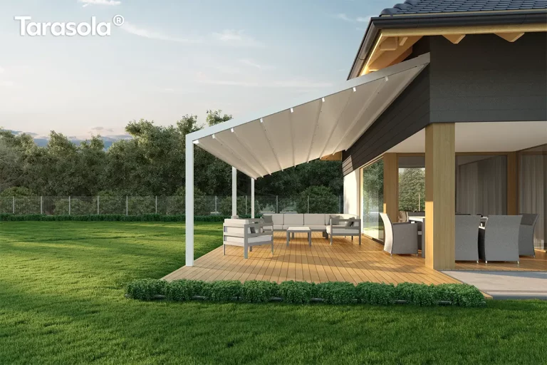 Biała pergola tkaninowa Tarasola Light w wersji przyściennej, zamontowana na jasnym drewnianym tarasie przy nowoczesnym domu o grafitowej elewacji z drewnianymi akcentami konstrukcyjnymi. Zadaszenie pergoli jest w pełni rozłożone, prezentując jasny, trójwarstwowy materiał Opatex o lekko pofalowanej strukturze, który chroni strefę wypoczynkową przed słońcem. Konstrukcja opiera się na smukłych, białych profilach aluminiowych, a pod tkaniną widoczne są poprzeczne belki zintegrowane z punktowym oświetleniem LED. Na tarasie znajduje się narożna, jasnoszara sofa z niskim stolikiem oraz osobny fotel, a w głębi, pod zadaszoną częścią budynku, widoczny jest duży stół z szarymi krzesłami ogrodowymi. Otoczenie stanowi intensywnie zielony trawnik z pasem niskich krzewów ozdobnych przy krawędzi desek oraz gęsta ściana drzew liściastych w tle, pod jasnym, popołudniowym niebem.