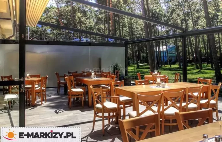 Widok z wnętrza dużej pergoli restauracyjnej Moderno o czarnej, aluminiowej konstrukcji, oszklonej przesuwnymi panelami bocznymi, które otwierają przestrzeń na otaczający las sosnowy. Wewnątrz ogródka gastronomicznego na szarej podłodze tarasowej ustawione są liczne drewniane stoliki z pasującymi krzesłami wyposażonymi w jasne poduszki. Zadaszenie pergoli jest całkowicie odsłonięte, co pozwala na pełny dostęp światła dziennego do wnętrza, a jedna z bocznych ścian posiada zamontowaną pionową przesłonę materiałową w kolorze mlecznym, zapewniającą prywatność i ochronę przed słońcem.