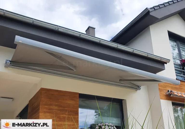 Nowoczesna markiza tarasowa w pełnej kasecie zamontowana na ścianie budynku mieszkalnego nad oknem tarasowym. Konstrukcja markizy oraz jej poszycie są w jednolitym, jasnoszarym kolorze, co nadaje całości minimalistyczny i elegancki wygląd. Ramiona przegubowe markizy są częściowo rozłożone, podtrzymując napiętą tkaninę osłony, która tworzy zadaszenie nad częścią elewacji. Zadaszenie zamontowane jest bezpośrednio pod ciemnoszarym podbitkiem dachu, w pobliżu metalowej rynny odpływowej. Elewacja domu łączy biały tynk z dekoracyjnymi elementami o strukturze naturalnego drewna w ciepłym, brązowym odcieniu, widocznymi na filarze oraz fragmencie ściany po prawej stronie. W dolnej części kadru widoczne są ozdobne trawy ogrodowe oraz fragmenty kwiatów w donicy stojącej na parapecie. Po prawej stronie, w tle, znajduje się drugie okno z częściowo widoczną mniejszą markizą w pomarańczowo-białe pasy. Całość kompozycji prezentuje produkt w naturalnym, nowoczesnym otoczeniu architektury domowej w jasny, lekko zachmurzony dzień.