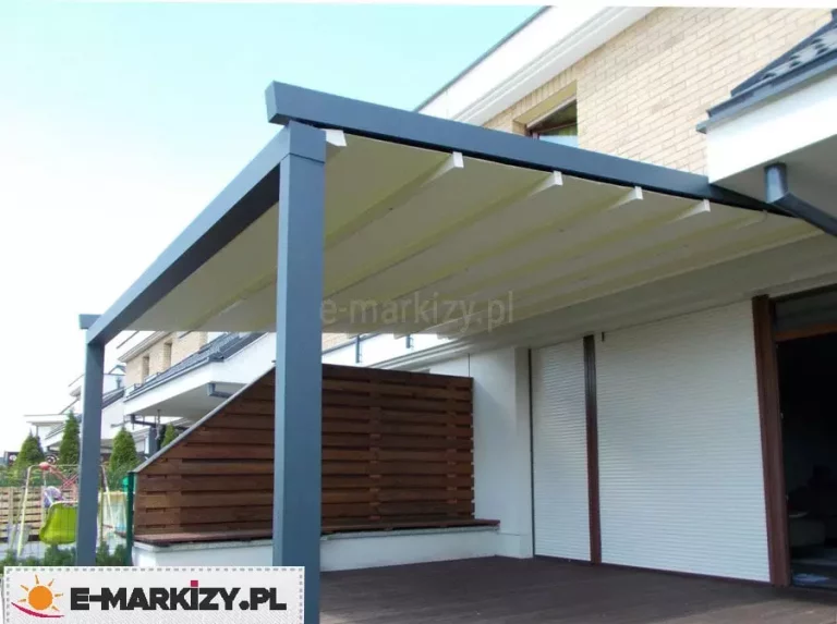 Zdjęcie przedstawia realizację tarasową, której centralnym punktem jest pergola przyścienna Moderno zamontowana do elewacji piętrowego budynku wykonanego z jasnej cegły oraz białego tynku. Konstrukcja pergoli ma kolor ciemnego antacytu i opiera się na solidnych, kwadratowych profilach bocznych. Zadaszenie składa się z rozsuwanej tkaniny w kolorze kremowym lub złamanej bieli, która jest rozpięta na poprzecznych belkach, tworząc estetyczne fałdy materiału chroniące przed słońcem. Pergola jest zamontowana pod rynną i okapem budynku, osłaniając przestronny taras wyłożony deskami kompozytowymi w kolorze ciemnego brązu. Na tarasie widoczna jest drewniana ściana działowa o schodkowym kształcie, pełniąca funkcję osłony od sąsiedniej posesji. W tle widać fragmenty innych budynków mieszkalnych, zielone krzewy oraz elementy placu zabaw z huśtawką. Elewacja budynku pod pergolą wyposażona jest w białe rolety zewnętrzne zasłaniające okna i drzwi tarasowe. Całość kompozycji ukazana jest w pełnym świetle dziennym przy bezchmurnym niebie.