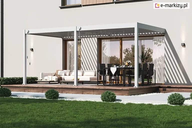 Dwumodułowa pergola lamelowa Marlo w kolorze białym, zamontowana do jasnej elewacji piętrowego budynku mieszkalnego. Konstrukcja zadaszenia wspiera się na trzech pionowych słupach o kwadratowym przekroju, które zostały osadzone na podwyższonym tarasie wykonanym z drewna w ciepłym odcieniu brązu. Każdy z dwóch modułów posiada oddzielny mechanizm korbowy do ręcznej regulacji kąta nachylenia lameli, widoczny przy słupach. Lamele w obu sekcjach są ustawione pod kątem, co powoduje powstawanie charakterystycznych, pasowych cieni na elewacji domu. Pod pergolą zaaranżowano dwie strefy: po lewej stronie znajduje się jasny narożnik wypoczynkowy z poduszkami i stolikiem kawowym, natomiast po prawej stronie ustawiono duży stół jadalniany z czarnymi krzesłami. W tle widoczne są duże przeszklenia tarasowe, a na ścianie budynku zamontowano dwa nowoczesne kinkiety emitujące światło góra-dół. Przed tarasem rozpościera się zadbany trawnik z równo przyciętymi, kulistymi krzewami ozdobnymi, oddzielony od konstrukcji pasem białego żwiru.