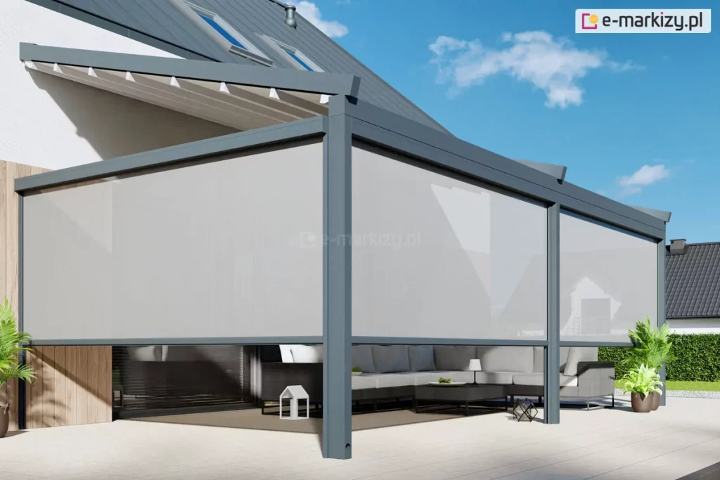 Wizualizacja przedstawiająca nowoczesną architekturę tarasową, której głównym elementem jest pergola przyścienna Moderno zamontowana do elewacji budynku o jasnym tynku. Konstrukcja pergoli została wykonana z solidnych profili aluminiowych w kolorze antracytowym, a jej zadaszenie stanowi biały, rozkładany dach tkaninowy wsparty na poprzecznych belkach. Na wszystkich otwartych bokach pergoli zainstalowano materiałowe rolety zewnętrzne typu Refleksol w kolorze jasnoszarym, które są częściowo rozwinięte, przesłaniając górną i środkową część przestrzeni wypoczynkowej. Pod zadaszeniem znajduje się zestaw mebli ogrodowych, w tym duża, narożna sofa w kolorze ciemnoszarym z jasnymi poduszkami oraz niski stolik kawowy, na którym umieszczono drobne dekoracje. Podłoże tarasu wyłożone jest dużymi, jasnymi płytami o matowym wykończeniu, a fragment ściany budynku wykończono pionowymi lamelami drewnianymi. W tle widoczny jest fragment dachu budynku z oknami połaciowymi oraz błękitne niebo z delikatnymi chmurami, a po bokach kompozycję dopełniają rośliny w donicach oraz zielony żywopłot.