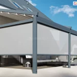Wizualizacja przedstawiająca nowoczesną architekturę tarasową, której głównym elementem jest pergola przyścienna Moderno zamontowana do elewacji budynku o jasnym tynku. Konstrukcja pergoli została wykonana z solidnych profili aluminiowych w kolorze antracytowym, a jej zadaszenie stanowi biały, rozkładany dach tkaninowy wsparty na poprzecznych belkach. Na wszystkich otwartych bokach pergoli zainstalowano materiałowe rolety zewnętrzne typu Refleksol w kolorze jasnoszarym, które są częściowo rozwinięte, przesłaniając górną i środkową część przestrzeni wypoczynkowej. Pod zadaszeniem znajduje się zestaw mebli ogrodowych, w tym duża, narożna sofa w kolorze ciemnoszarym z jasnymi poduszkami oraz niski stolik kawowy, na którym umieszczono drobne dekoracje. Podłoże tarasu wyłożone jest dużymi, jasnymi płytami o matowym wykończeniu, a fragment ściany budynku wykończono pionowymi lamelami drewnianymi. W tle widoczny jest fragment dachu budynku z oknami połaciowymi oraz błękitne niebo z delikatnymi chmurami, a po bokach kompozycję dopełniają rośliny w donicach oraz zielony żywopłot.