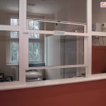 Biała zabudowa okienna typu recepcja, wykonaną z profili aluminiowych z przeszkleniem. Centralnym elementem jest poziome, przesuwne okienko podawcze z napisem "RECEPTION" umieszczonym na ramie, zabezpieczone małym zamkiem punktowym w górnej części. Konstrukcja osadzona jest na ścianie o koralowym lub ceglastym kolorze, wykończonej białym parapetem. Przez szyby widać wnętrze biura z ciemnym fotelem biurowym, szafami dokumentowymi oraz klasycznym, białym oknem w tle, przez które prześwitują drzewa.