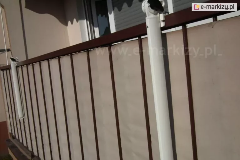 Zbliżenie na system montażowy ramion jakie posiada markiza balkonowo-poręczowa, zainstalowanych na balustradzie tarasu. Zdjęcie prezentuje dwa białe, pionowe ramiona mocujące, które są przytwierdzone do ciemnobrązowej barierki balkonowej wykonanej z metalowych, pionowych szczebli. Ramiona poręczowe zwisają pionowo w dół wzdłuż balustrady balkonu, w górnej - mocowanej do barierki części - posiadają mechanizm regulacyjny z czarnymi elementami blokującymi u góry, umożliwiający stabilne osadzenie konstrukcji między poręczą a podłożem lub górną płytą balkonu. Za barierką widoczna jest gładka osłona balkonowa w kolorze beżowym, która zapewnia prywatność i ochronę przed wiatrem. W tle znajduje się jasna elewacja budynku oraz okno z częściowo opuszczoną białą roletą zewnętrzną. Konstrukcja markizy balkonowo-poręczowej charakteryzuje się minimalistycznym designem i solidnym wykonaniem z lakierowanego proszkowo aluminium, co gwarantuje odporność na warunki atmosferyczne. Padające z boku światło słoneczne tworzy wyraźne cienie na osłonie, podkreślając geometrię szczebli balustrady oraz detale techniczne uchwytów montażowych.