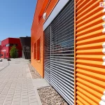 Fasada nowoczesnego budynku przemysłowo-usługowego o elewacji z intensywnie pomarańczowych, poziomo ryflowanych blach trapezowych. Po prawej stronie kadru znajduje się duże okno osłonięte zewnętrzną żaluzją fasadową z lamelami w kolorze srebrnym. Lamele są poziome, typu C, zamontowane w prowadnicach bocznych i ustawione w pozycji lekko uchylonej, umożliwiając częściowy wgląd do wnętrza. Nad żaluzją zamontowana jest maskownica kasety w kolorze dopasowanym do lameli. Na powierzchni blach elewacyjnych widoczny jest nadruk w kolorze białym, stanowiący część napisu lub logotypu. Przy ścianie budynku ułożone są drobne kamienie ozdobne jako element aranżacji przestrzeni przyokiennej. Chodnik wykonany z prostokątnych płyt betonowych biegnie wzdłuż budynku, prowadząc w kierunku kolejnych obiektów. W tle widoczne są kolejne budynki o podobnej bryle, utrzymane w czerwonej kolorystyce. Na jednym z nich widnieje biały napis "świetlica". Na ulicy zaparkowanych jest kilka samochodów osobowych. Po lewej stronie zdjęcia znajdują się nowoczesne latarnie uliczne oraz kuliste elementy betonowe pełniące funkcję małej architektury. Całość przedstawia uporządkowaną przestrzeń miejską o nowoczesnym charakterze.