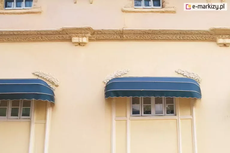 Zdjęcie przedstawia fragment klasycznej fasady budynku w kolorze piaskowym, ozdobionej bogatymi detalami architektonicznymi, na której zamontowano dwie eleganckie markizy koszowe o wydłużonym, prostokątnym profilu frontowym. Markizy posiadają poszycie w kolorze głębokiego granatu, które jest podzielone poziomymi, cienkimi białymi liniami, co nadaje im luksusowy charakter. Dolna krawędź każdej osłony wykończona jest falowaną falbaną z wyraźną, jasną lamówką. Markizy są umieszczone bezpośrednio nad oknami o białych ramach, a nad każdą z nich znajduje się dekoracyjny sztukatorski element w kształcie liścia lub pióra. Elewacja powyżej markiz posiada wydatny, ozdobny gzyms wsparty na konsolach, co sugeruje zabytkowy lub stylizowany charakter obiektu. Całość kompozycji jest symetryczna i utrzymana w stonowanej, eleganckiej kolorystyce, a jasne oświetlenie podkreśla teksturę tynku oraz detale wykończenia markiz.