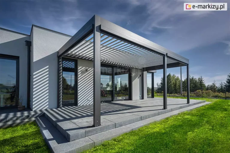 Nowoczesny budynek utrzymany w tonacjach szarości z przestronnym tarasem z płyt betonowych przy zielonym trawniku osłoniętym pergolą z dachem lamelowym SB400. Antracytowa konstrukcja pergoli wpisuje się w kolorystykę ram dużych okien w budynku. W tle widoczne drzewa oraz niebieskie niebo.