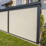 Dwa refleksole Ziiip 120 Selt zamocowane do konstrukcji pergoli, w kolorze konstrukcji antracytowym, dopasowanym do barw zadaszenia. Beżowa tkanina chroni przed słońcem i niepożądanymi spojrzeniami. Przed roletami widoczny trawnik a obok pergoli kwietnik z zielonymi roślinami.