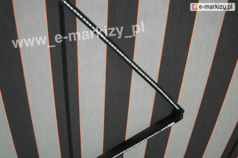 Oświetlenie LED do markiz zamocowane do ramienia markizy Palladio o konstrukcji w kolorze srebrzysty grafit z rozwiniętym poszyciem w pasy szare, grafitowe oraz czerwone. Włączone oświetlenie posiada zimną barwę światła.