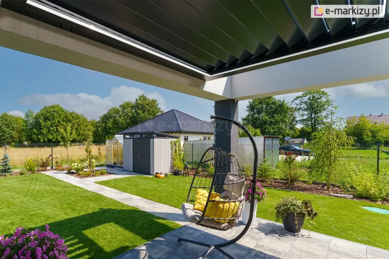 Zdjęcie prezentuje perspektywę od strony tarasu na ogród, ukazując system bioklimatycznego dachu lamelowego SB400R w pozycji zamkniętej. Aluminiowe lamele w kolorze antracytowym tworzą szczelną i gładką płaszczyznę zadaszenia, chroniącą przed warunkami atmosferycznymi. Pod krawędzią ramy konstrukcyjnej zamontowano liniowe oświetlenie LED, które emituje jasne światło, doświetlając strefę wypoczynkową. Konstrukcja dachu zamocowana jest w betonowej białej opasce, która wsparta jest na ciemnoszarym słupie nośnym. Posadzka tarasu wykonana jest z jasnoszarych płyt betonowych. Na pierwszym planie widoczny jest wiszący fotel ogrodowy z szarymi poduszkami, a w donicach umieszczono kwitnące fioletowe kwiaty. W tle rozpościera się widok na zadbany trawnik, szary domek narzędziowy o nowoczesnej bryle oraz liczne nasadzenia roślinne pod błękitnym niebem z rozproszonymi chmurami.