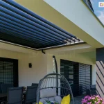 Nowoczesny budynek mieszkalny z tarasem wyposażonym w dach lamelowy SB400R Aluprof. Aluminiowe lamele dachowe w kolorze antracytowym są ustawione w pozycji otwartej (pionowej), co zapewnia maksymalny dopływ światła dziennego i pełną wentylację strefy wypoczynkowej. Rama zadaszenia posiada zintegrowane oświetlenie liniowe LED, które jest widoczne wzdłuż wewnętrznych krawędzi konstrukcji. Pod pergolą znajduje się komplet mebli tarasowych: ciemny stół z krzesłami oraz wiszący fotel ogrodowy z szarymi poduszkami. Jasna elewacja domu kontrastuje z ciemnymi słupami nośnymi pergoli oraz antracytowymi żaluzjami fasadowymi zamontowanymi w oknach. Na tarasie wyłożonym płytami betonowymi ustawiono donice z fioletowymi kwiatami, a w tle widoczna jest otwarta przestrzeń z zieloną roślinnością pod lekko zachmurzonym niebem.