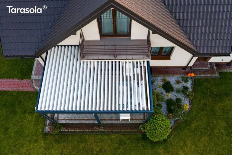 Widok z lotu ptaka przedstawia nowoczesną pergolę tarasową Essential Tarasola zamontowaną przy ścianie piętrowego domu jednorodzinnego. Konstrukcja zadaszenia opiera się na ciemnoszarym, antracytowym stelażu wykonanym z metalowych profili, który przylega bezpośrednio do elewacji budynku pod balkonem znajdującym się na wyższej kondygnacji. Dach lamelowy Tarasola Essential składa się z białych, równolegle ułożonych lameli, które są w tej chwili częściowo otwarte, tworząc pasy światła i cienia na powierzchni tarasu pod nimi. Pod zadaszeniem widoczna jest przestrzeń wypoczynkowa z jasnymi meblami ogrodowymi ustawionymi na nawierzchni z szarej kostki brukowej, która jest wykończona ozdobną obwódką z ciemniejszej kostki. Dom posiada białą elewację oraz dach pokryty ciemną dachówką o falistym profilu. Nad pergolą znajduje się balkon z metalową, minimalistyczną balustradą w kolorze brązowym oraz przeszklone drzwi balkonowe osadzone w drewnianej stolarce. Otoczenie budynku to zadbany, zielony trawnik, przez który przebiega ścieżka z czerwonej kostki brukowej prowadząca do tarasu. Po prawej stronie pergoli znajduje się ozdobna rabata ogrodowa wysypana drobnym, jasnym żwirem, na której posadzono różnorodne rośliny ozdobne, w tym niskie krzewy iglaste i liściaste oraz większy, gęsty krzew przy samym narożniku konstrukcji. Na krawędzi opaski wokół domu, niedaleko wejścia, widoczne są dwie pomarańczowe dynie stanowiące element dekoracyjny. Całość kompozycji ukazuje funkcjonalne połączenie nowoczesnej architektury ogrodowej z klasyczną bryłą domu.