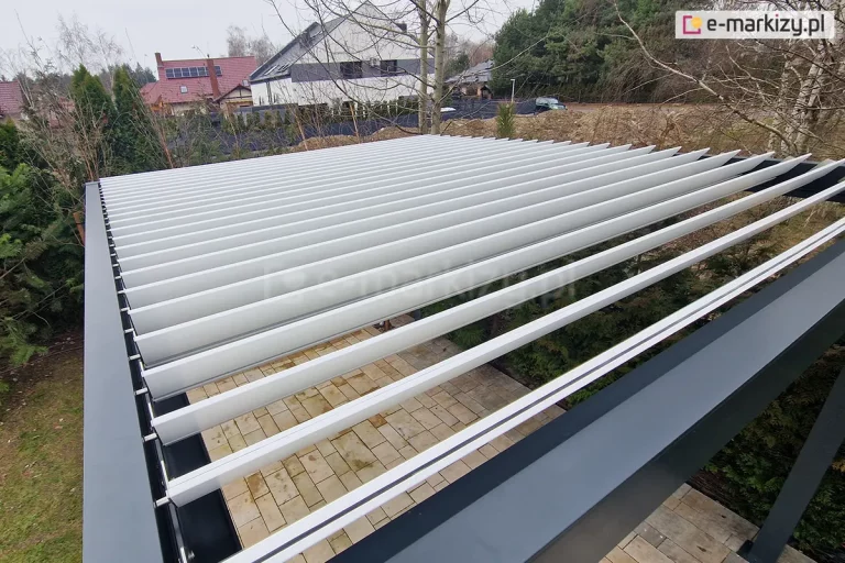 Widok z góry na dach pergoli bioklimatycznej Aluprof z lamelami ustawionymi w pozycji półotwartej (pod kątem). Aluminiowe lamele w kolorze jasnoszarym są zamocowane w solidnej, szerokiej ramie o kolorze antracytowym. Konstrukcja ukazuje mechanizm obrotowy oraz wewnętrzne koryta systemu odprowadzania wody deszczowej. Przez szczeliny między lamelami widoczny jest fragment tarasu wyłożonego jasną kostką brukową. W tle rozpościera się widok na ogród z gęstymi iglakami, brzozami oraz nowoczesną zabudowę jednorodzinną z dachami krytymi dachówką i panelami fotowoltaicznymi pod zachmurzonym niebem.