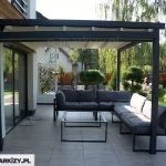 Zdjęcie prezentuje nowoczesną pergolę tarasową Luxo w zabudowie przyściennej, zainstalowaną na nowoczesnym budynku mieszkalnym. Konstrukcja wykonana jest z ekstrudowanego aluminium w kolorze antracytowym, składająca się z solidnej ramy zamocowanej do elewacji oraz dwóch zewnętrznych słupów wsporczych o kwadratowym przekroju. Zadaszenie stanowi ruchoma, segmentowa tkanina w kolorze kremowym lub jasnobeżowym, która w aktualnej konfiguracji jest całkowicie rozłożona, zapewniając cień nad strefą wypoczynkową. Pod pergolą znajduje się zestaw mebli ogrodowych składający się z modułowej sofy narożnej z szarymi poduszkami oraz minimalistycznego stolika kawowego o lekkiej, metalowej konstrukcji. Podłoże tarasu wyłożone jest dużymi płytami gresowymi w kolorze szarym. W tle widoczny jest zadbany ogród z niskimi krzewami formowanymi w kule, biała ażurowa kratka ogrodowa z roślinnością pnącą oraz drewniane ogrodzenie posesji. Całość przedstawia funkcjonalne wykorzystanie przestrzeni tarasowej dzięki systemowi otwieranego zadaszenia.
