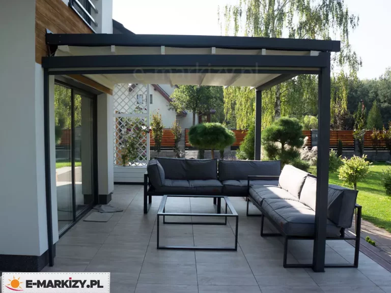 Zdjęcie prezentuje nowoczesną pergolę tarasową Luxo w zabudowie przyściennej, zainstalowaną na nowoczesnym budynku mieszkalnym. Konstrukcja wykonana jest z ekstrudowanego aluminium w kolorze antracytowym, składająca się z solidnej ramy zamocowanej do elewacji oraz dwóch zewnętrznych słupów wsporczych o kwadratowym przekroju. Zadaszenie stanowi ruchoma, segmentowa tkanina w kolorze kremowym lub jasnobeżowym, która w aktualnej konfiguracji jest całkowicie rozłożona, zapewniając cień nad strefą wypoczynkową. Pod pergolą znajduje się zestaw mebli ogrodowych składający się z modułowej sofy narożnej z szarymi poduszkami oraz minimalistycznego stolika kawowego o lekkiej, metalowej konstrukcji. Podłoże tarasu wyłożone jest dużymi płytami gresowymi w kolorze szarym. W tle widoczny jest zadbany ogród z niskimi krzewami formowanymi w kule, biała ażurowa kratka ogrodowa z roślinnością pnącą oraz drewniane ogrodzenie posesji. Całość przedstawia funkcjonalne wykorzystanie przestrzeni tarasowej dzięki systemowi otwieranego zadaszenia.