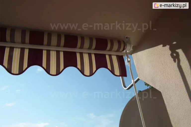 Fotografia prezentuje zbliżenie na mechanizm mocujący oraz detale wykończenia produktu, jakim jest markiza balkonowa poręczowa. Konstrukcja została zamontowana bezpośrednio do sufitu balkonu za pomocą białego, metalowego uchwytu, który trzyma rurę nawojową z tkaniną. Markiza balkonowa poręczowa posiada materiał w klasyczne pionowe pasy o barwie bordowej, beżowej i białej, zakończony ozdobną falbaną z czerwoną lamówką. Po prawej stronie widoczny jest mechanizm korbowy z białym uchem, do którego wkłada się drążek służący do ręcznego rozwijania i zwijania osłony. Zdjęcie wykonano z perspektywy od dołu, ukazując jasne, błękitne niebo w tle oraz fragment elewacji budynku o chropowatej strukturze tynku w kolorze piaskowym. Cień rzucany przez elementy konstrukcyjne na ścianę sugeruje ekspozycję na silne światło słoneczne, co podkreśla funkcję ochronną markizy.