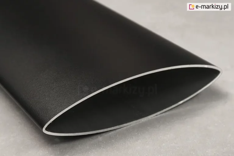 Zbliżenie na pojedynczy profil aluminiowy stanowiący element panelu żaluzjowego Louvre, prezentujący jego techniczny przekrój. Element ma charakterystyczny, aerodynamiczny kształt soczewki lub wydłużonej elipsy o ostrych krawędziach bocznych. Powierzchnia profilu jest wykończona matową, lekko ziarnistą strukturą w kolorze głębokiego antracytu lub czerni. Na krawędzi cięcia widoczna jest naturalna, srebrzysta barwa aluminium, co podkreśla grubość ścianki profilu. Przedmiot spoczywa na jasnoszarym, jednolitym podłożu o delikatnie porowatej fakturze, przypominającej beton lub kamień. Oświetlenie jest miękkie i rozproszone, co pozwala na dokładne zaobserwowanie satynowego połysku na obłej powierzchni detalu oraz precyzji jego wykonania.