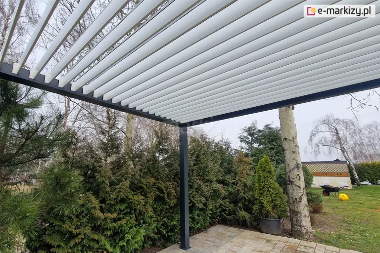 Widok od dołu na zadaszenie pergoli bioklimatycznej Sb350 Aluprof z lamelami ustawionymi w pozycji półotwartej. Aluminiowe lamele w kolorze jasnoszarym są zamontowane w czarnej (antracytowej) ramie, co pozwala na regulację dostępu światła i wentylację tarasu. Konstrukcja opiera się na słupie o kwadratowym przekroju, przytwierdzonym do podłoża z jasnych płyt betonowych. W tle widoczny jest ogród z gęstą roślinnością: ciemnozielonymi iglakami, wysoką brzozą o jasnej korze oraz zadbanym trawnikiem, na którym leży żółta piłka i taczka ogrodowa. Zdjęcie wykonane w świetle dziennym przy zachmurzonym niebie, prezentujące techniczne aspekty działania dachu lamelowego.