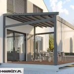 Nowoczesna pergola przyścienna Moderno o konstrukcji z ciemnoantracytowych profili aluminiowych, zamontowana na podwyższonym betonowym tarasie wyłożonym drewnianymi deskami. Zadaszenie stanowi ruchoma tkanina techniczna w kolorze ciemnoszarym, ułożona w równe segmenty na prowadnicach, co zapewnia osłonę przed słońcem i deszczem. Jedna ze ścian bocznych została wyposażona w pionową przesłonę typu refleksol w jasnym, półprzezroczystym kolorze, która chroni wnętrze przed bocznym nasłonecznieniem i wiatrem. Pod konstrukcją ustawiono zestaw mebli jadalnianych składający się z prostokątnego stołu oraz nowoczesnych krzeseł, a całość przylega do białej elewacji budynku z dużymi, przeszklonymi drzwiami tarasowymi i oknami wyposażonymi w żaluzje fasadowe.