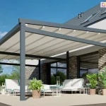 Nowoczesna pergola tarasowa Moderno, charakteryzująca się solidną konstrukcją z profili aluminiowych w kolorze antracytowym. Zadaszenie stanowi jasny, segmentowy materiał tkaninowy, który jest w pełni rozwinięty i zamontowany na prowadnicach pod lekkim kątem, co ułatwia odpływ wody. Konstrukcja przylega do elewacji budynku wykończonej jasną cegłą klinkierową z dużymi przeszkleniami. Na jasnym tarasie pod pergolą zaaranżowano strefę wypoczynkową z białymi sofami o błękitnych lamówkach, białymi stolikami kawowymi oraz roślinami w donicach, co tworzy spójną i elegancką przestrzeń rekreacyjną.