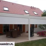 Zdjęcie przedstawia pergolę rzymską Moderno w wersji przyściennej, wyposażoną w pionowe osłony przeciwsłoneczne typu Zip. Konstrukcja pergoli oraz kasety osłon bocznych są w kolorze kremowym lub jasnobeżowym, co koresponduje z jasną elewacją budynku. Dwie przednie rolety są częściowo rozwinięte, przesłaniając górną część prześwitu między profilami pionowymi. Pod zadaszeniem znajduje się strefa wypoczynkowa z brązowymi meblami technorattanowymi oraz grillem. Budynek posiada dach pokryty czerwoną dachówką z trzema oknami połaciowymi, a okna parterowe zdobią drewniane, brązowe okiennice. Na tarasie oraz przy jego krawędzi ustawiono liczne donice z kwitnącymi, różowymi kwiatami, a całość graniczy bezpośrednio z trawnikiem.