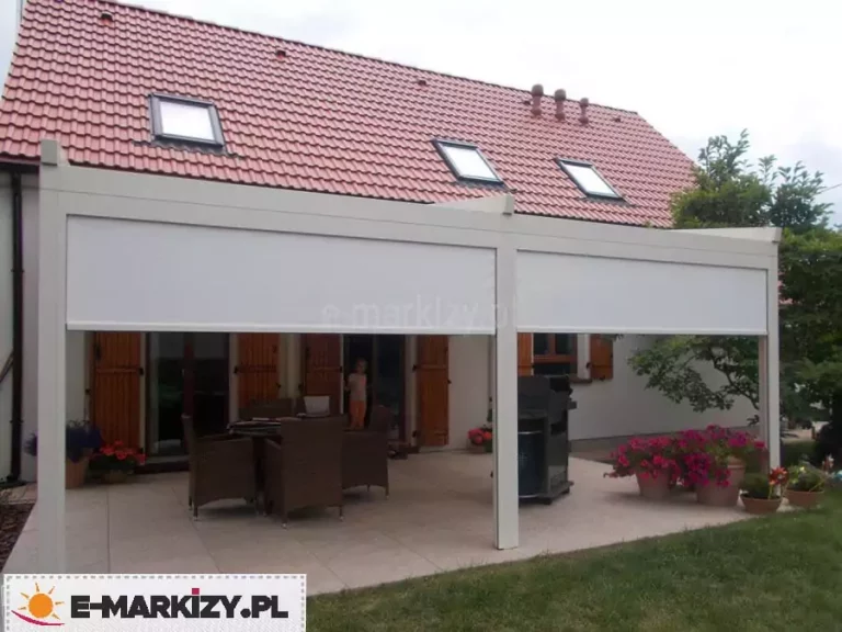 Zdjęcie przedstawia pergolę rzymską Moderno w wersji przyściennej, wyposażoną w pionowe osłony przeciwsłoneczne typu Zip. Konstrukcja pergoli oraz kasety osłon bocznych są w kolorze kremowym lub jasnobeżowym, co koresponduje z jasną elewacją budynku. Dwie przednie rolety są częściowo rozwinięte, przesłaniając górną część prześwitu między profilami pionowymi. Pod zadaszeniem znajduje się strefa wypoczynkowa z brązowymi meblami technorattanowymi oraz grillem. Budynek posiada dach pokryty czerwoną dachówką z trzema oknami połaciowymi, a okna parterowe zdobią drewniane, brązowe okiennice. Na tarasie oraz przy jego krawędzi ustawiono liczne donice z kwitnącymi, różowymi kwiatami, a całość graniczy bezpośrednio z trawnikiem.