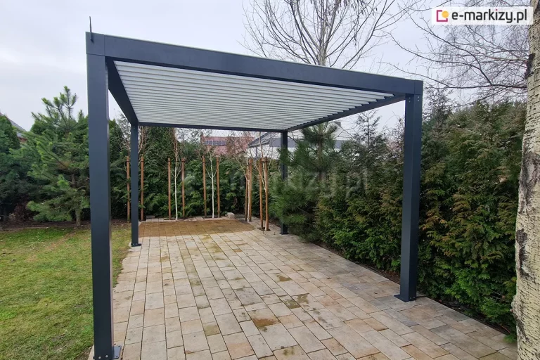Szerokie ujęcie nowoczesnej, wolnostojącej pergoli bioklimatycznej SB350 Selt zainstalowanej w ogrodzie na podłożu z jasnej kostki brukowej. Konstrukcja wykonana jest z profili aluminiowych malowanych proszkowo na kolor antracytowy, a jej dach stanowią białe, ruchome lamele, które na zdjęciu są całkowicie zamknięte, tworząc szczelną i płaską płaszczyznę. Pergola wsparta jest na czterech solidnych słupach o kwadratowym przekroju, przy czym jeden z nich znajduje się w bliskim sąsiedztwie pnia brzozy o charakterystycznej białej korze. W otoczeniu konstrukcji widoczna jest gęsta i zróżnicowana roślinność ogrodowa: wysokie żywopłoty z tui, młode drzewa liściaste wsparte na palikach oraz krzewy iglaste (sosny). Po lewej stronie kadru znajduje się fragment trawnika. W tle, ponad linią ogrodzenia i roślinności, widać dachy sąsiednich domów jednorodzinnych pod zachmurzonym, jasnym niebem. Całość prezentuje minimalistyczną architekturę ogrodową, która integruje się z naturalnym otoczeniem.