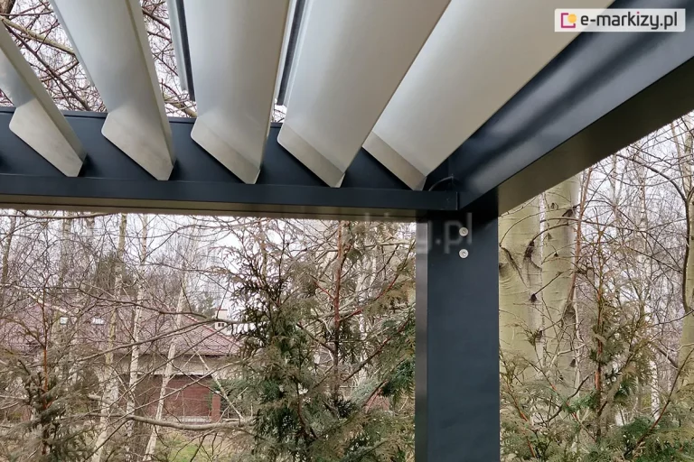 Zbliżenie na narożnik wewnętrzny pergoli bioklimatycznej SB 350 Aluprof, prezentujące system mocowania i pracy ruchomych lamel. Konstrukcja ramy oraz pionowy słup wsporczy wykonane są z aluminium lakierowanego proszkowo na kolor antracytowy. Białe, aluminiowe lamele dachowe o aerodynamicznym kształcie są zamontowane pod kątem (pozycja półotwarta), co ukazuje ich profil oraz sposób osadzenia w bocznej rurze napędowej. Na pionowym słupie widoczne są dwa srebrne punkty montażowe (śruby imbusowe). W prawym górnym rogu, przy łączeniu profili, widoczny jest czarny przewód elektryczny doprowadzający zasilanie do mechanizmu obrotowego lub systemu oświetlenia. Tło zdjęcia stanowią pnie i gałęzie brzóz oraz fragmenty krzewów iglastych, a w oddali widoczny jest zarys budynku mieszkalnego z czerwoną dachówką. Perspektywa od dołu podkreśla techniczną precyzję pasowania elementów konstrukcyjnych.