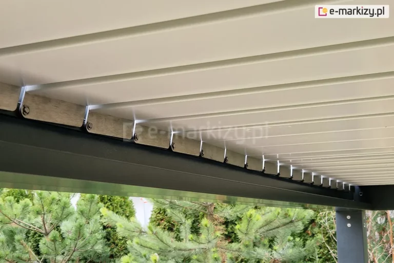 Zbliżenie od dołu na mechanizm obrotowy lamel w nowoczesnej pergoli bioklimatycznej SB350 Aluprof. Zdjęcie przedstawia białe, aluminiowe pióra (lamele) dachu, które są zamontowane do bocznej ramy konstrukcyjnej w kolorze antracytowym. Widoczne są stalowe elementy mocujące oraz system zawiasów, które umożliwiają regulację kąta nachylenia lamel w celu kontroli nasłonecznienia i wentylacji. W dolnej części kadru, pod konstrukcją, widoczne są korony drzew iglastych (sosny) oraz fragment pionowego słupa wsporczego pergoli z widocznymi śrubami montażowymi. Detal ten podkreśla precyzję wykonania ruchomych elementów zadaszenia oraz estetyczne połączenie kolorystyczne bieli lamel z ciemną ramą.