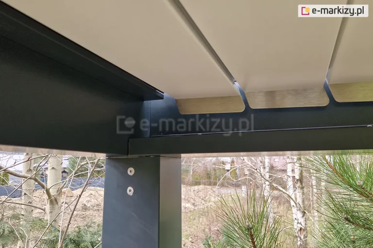 Techniczne zbliżenie na narożnik wewnętrzny pergoli bioklimatycznej Aluprof SB350, przedstawiające system mocowania dachu do słupa nośnego. Konstrukcja wykonana jest z aluminium w kolorze antracytowym, z widocznymi dwiema srebrnymi śrubami montażowymi na pionowym słupie. W górnej części widoczne są końce uchylnych lameli w kolorze białym ze srebrnymi, metalowymi nakładkami na krawędziach. Lamele są szczelnie zamknięte, a pod nimi widoczny jest mechanizm rynny odpływowej wewnątrz ramy. W tle, poza obrysem konstrukcji, widoczny jest ogród z iglakami (sosna), brzozami oraz zarys budynku mieszkalnego pod pochmurnym niebem.