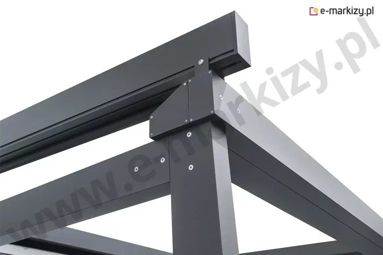 Zbliżenie na górny narożnik konstrukcji pergoli Solid Aluprof, wykonane z perspektywy dolnej, prezentujące systemowe łączenia profili oraz konstrukcję daszka ochronnego. Na zdjęciu widać punkt styku masywnego, pionowego słupa z belkami obwodowymi ramy o matowym, antracytowym wykończeniu. Newralgicznym elementem jest wieloczęściowa, trójkątna nakładka maskująca, która spaja elementy ramy i jest przytwierdzona za pomocą precyzyjnie rozmieszczonych śrub imbusowych ze stali nierdzewnej. Powyżej linii ramy głównej zamontowany jest wspornik w kształcie litery L, który podtrzymuje poziomy profil daszka ochronnego, osłaniającego mechanizm jezdny tkaniny. Całość konstrukcji charakteryzuje się surową, techniczną estetyką oraz wysoką jakością powłoki lakierniczej. Ujęcie to podkreśla solidność aluminiowych komponentów oraz dbałość o detale montażowe, które zapewniają stabilność i trwałość całego systemu w trudnych warunkach zewnętrznych.